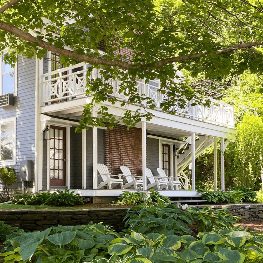 A two-story house with a porch, white chairs, and lush green plants.