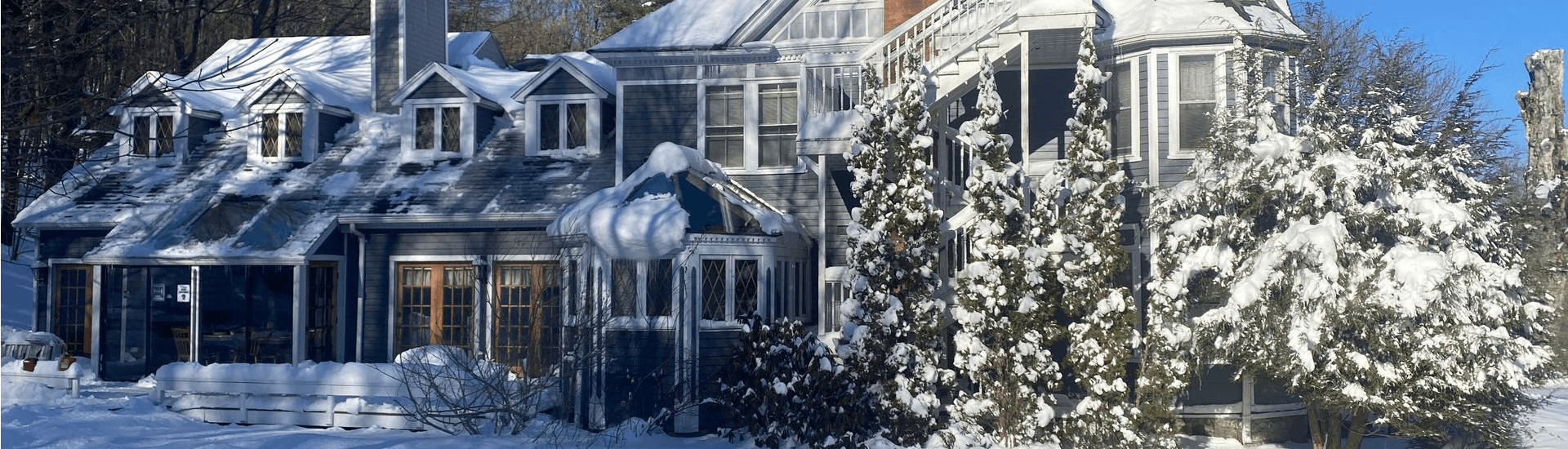 A snow-covered house with large windows and surrounding evergreen trees.