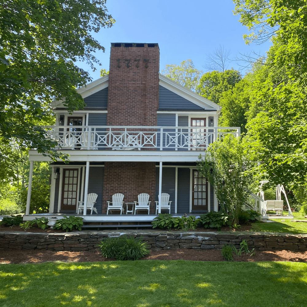 A two-story blue house with a brick chimney and white chairs on the porch, surrounded by greenery.