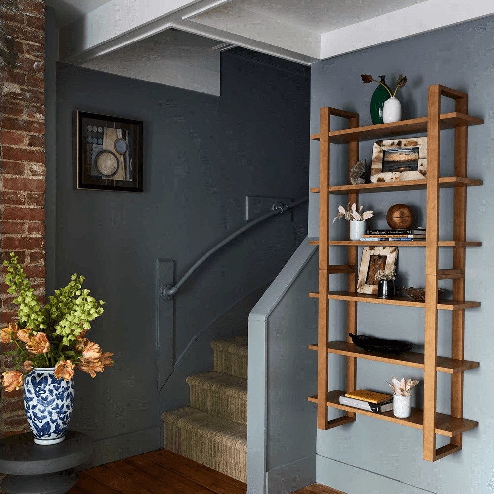 A wooden bookshelf with decorative items stands beside a staircase in a cozy, modern living space.