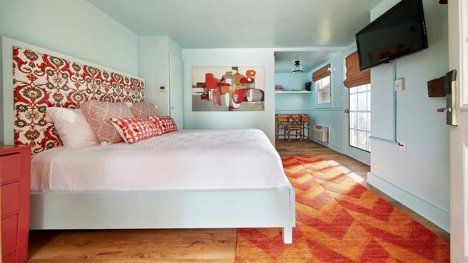 Small, colorful bedroom with patterned headboard, orange rug, and TV.