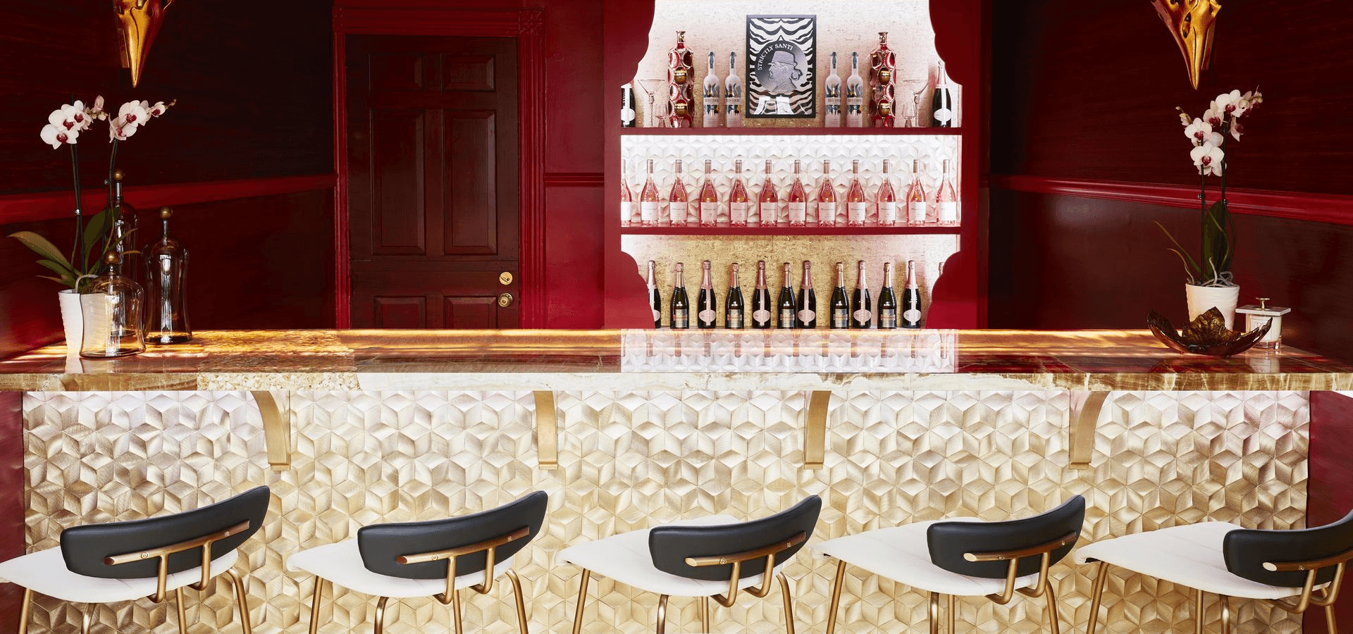 A sleek bar area featuring a golden textured surface, modern stools, and shelves lined with bottles against a rich red backdrop.