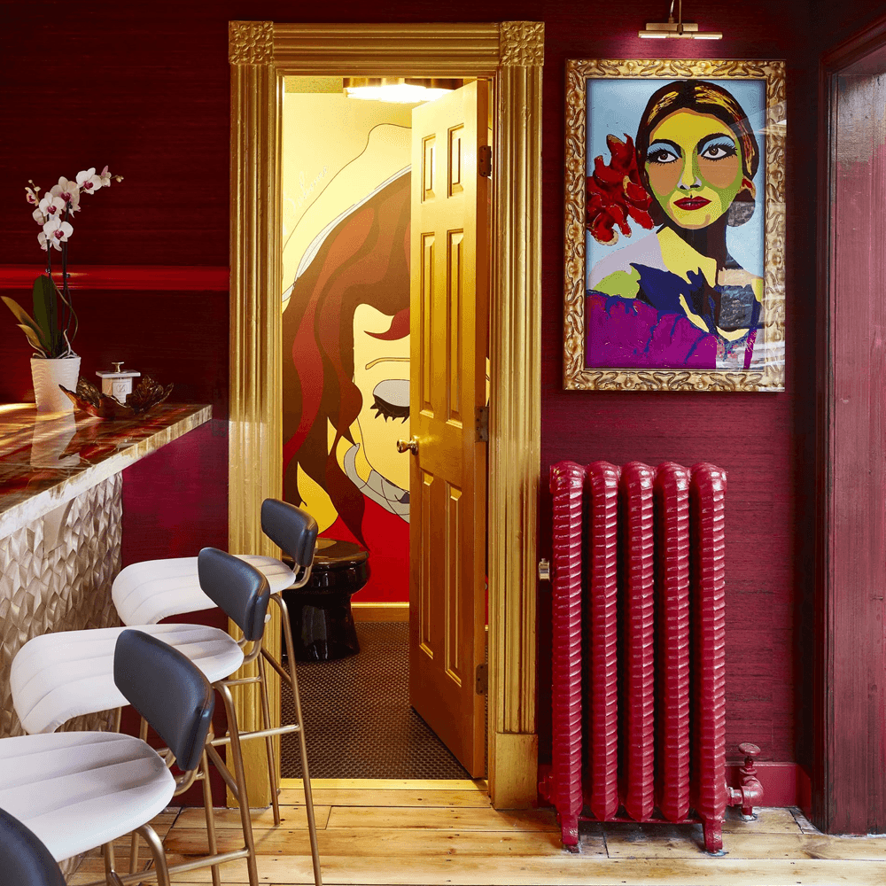 A colorful interior featuring a gold-framed painting, a deep red wall, a stylish bar with white and gold stools, and a doorway leading to a bathroom decorated with vibrant artwork.