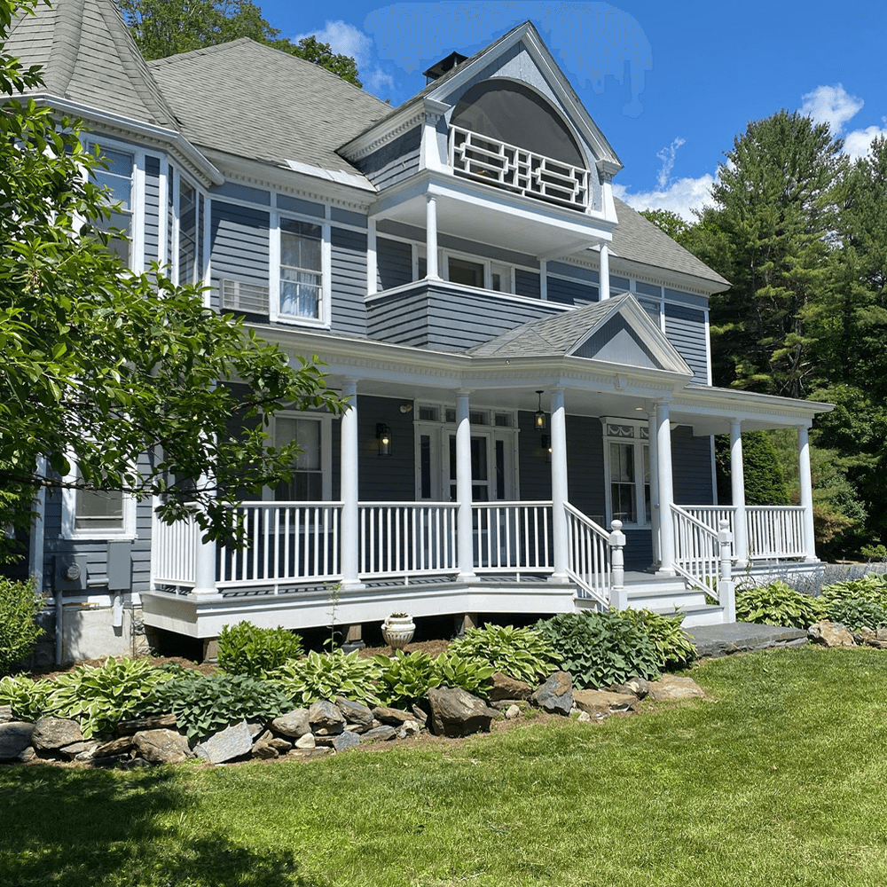 A large, blue Victorian-style house with a spacious porch surrounded by landscaped greenery.
