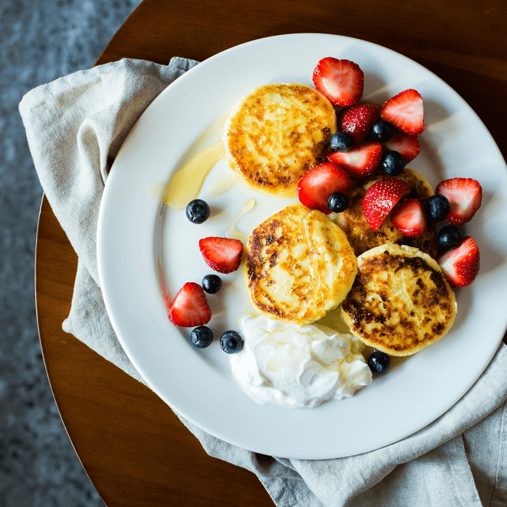 Fluffy pancakes topped with strawberries, blueberries, honey, and a dollop of whipped cream on a white plate.